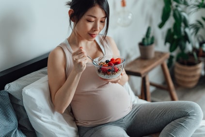 Pregnant woman eating a bowl of granola with yoghurt and mixed berries, sitting on bed
