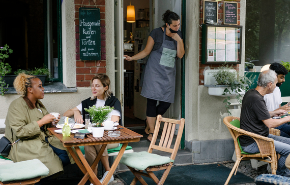 A busy restaurant façade with a waitress watching while people are sitting and eating outdoors.