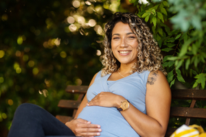 smiling young pregnant woman cradling her tummy while sitting outside on a park bench