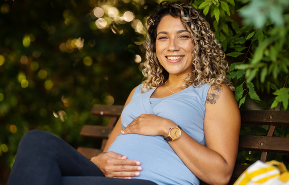 smiling pregnant woman cradling her tummy while sitting outside on a park bench