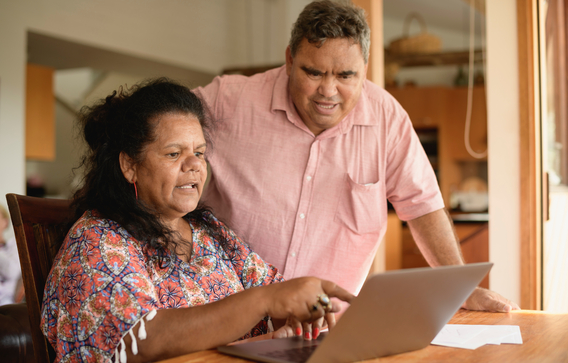 Aboriginal couple in their 50s at home using the internet, man leaning over woman and looking at laptop