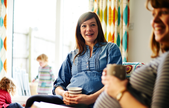 Two heavily pregnant women in lounge enjoying hot drinks and smiling while children play together in background.