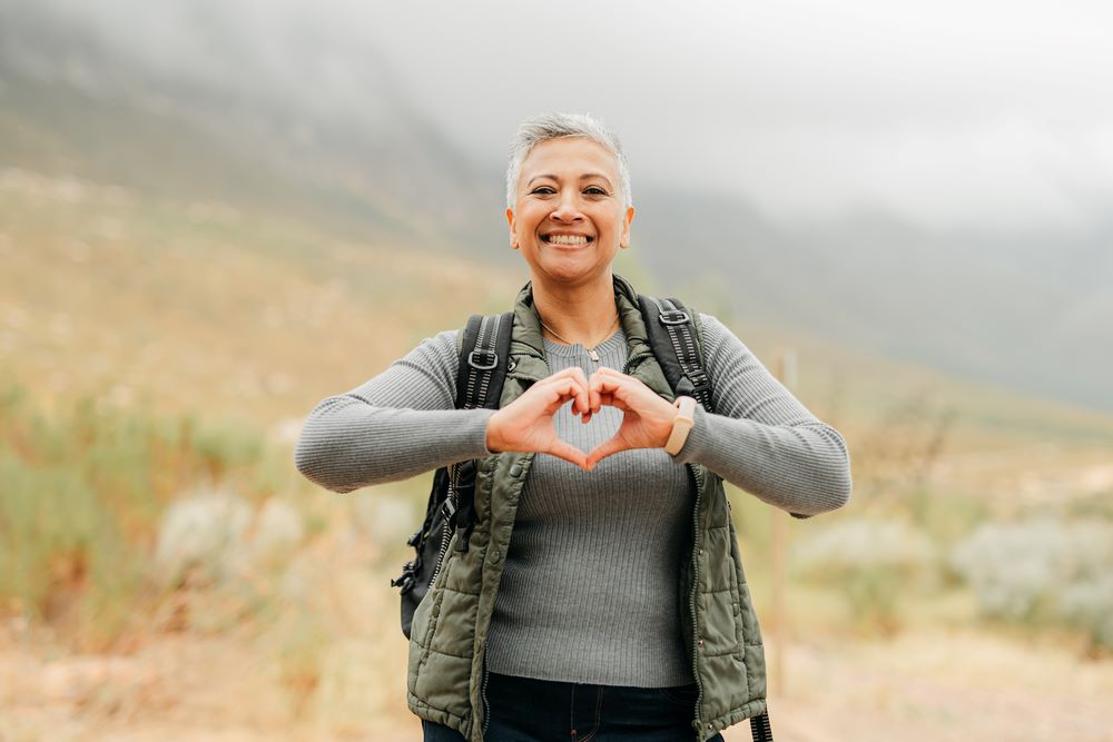 Woman standing outside in a field, looking at camera, making heart symbol with hands